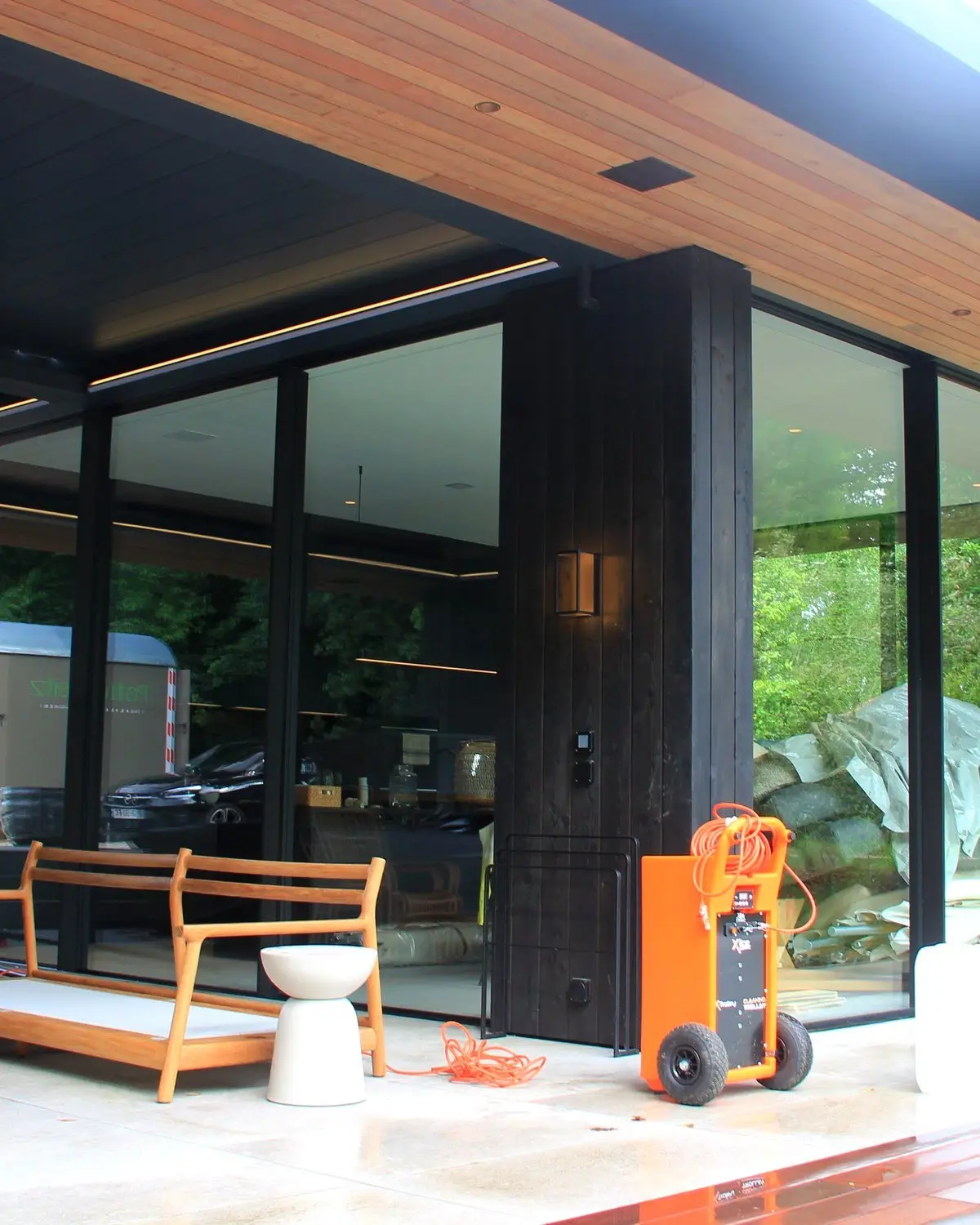 Terrasse d'un pavillon de piscine vitré, piliers en bois sombre, banc en bois et chariot à eau orange